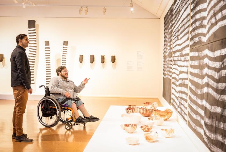 휠체어를 탄 남성이 아랄루엔 아츠 센터에서 미술 작품을 감상하고 있고 옆에 다른 남성이 서 있는 모습, 앨리스 스프링스, 노던테리토리 © 노던테리토리 관광청/헬렌 오르