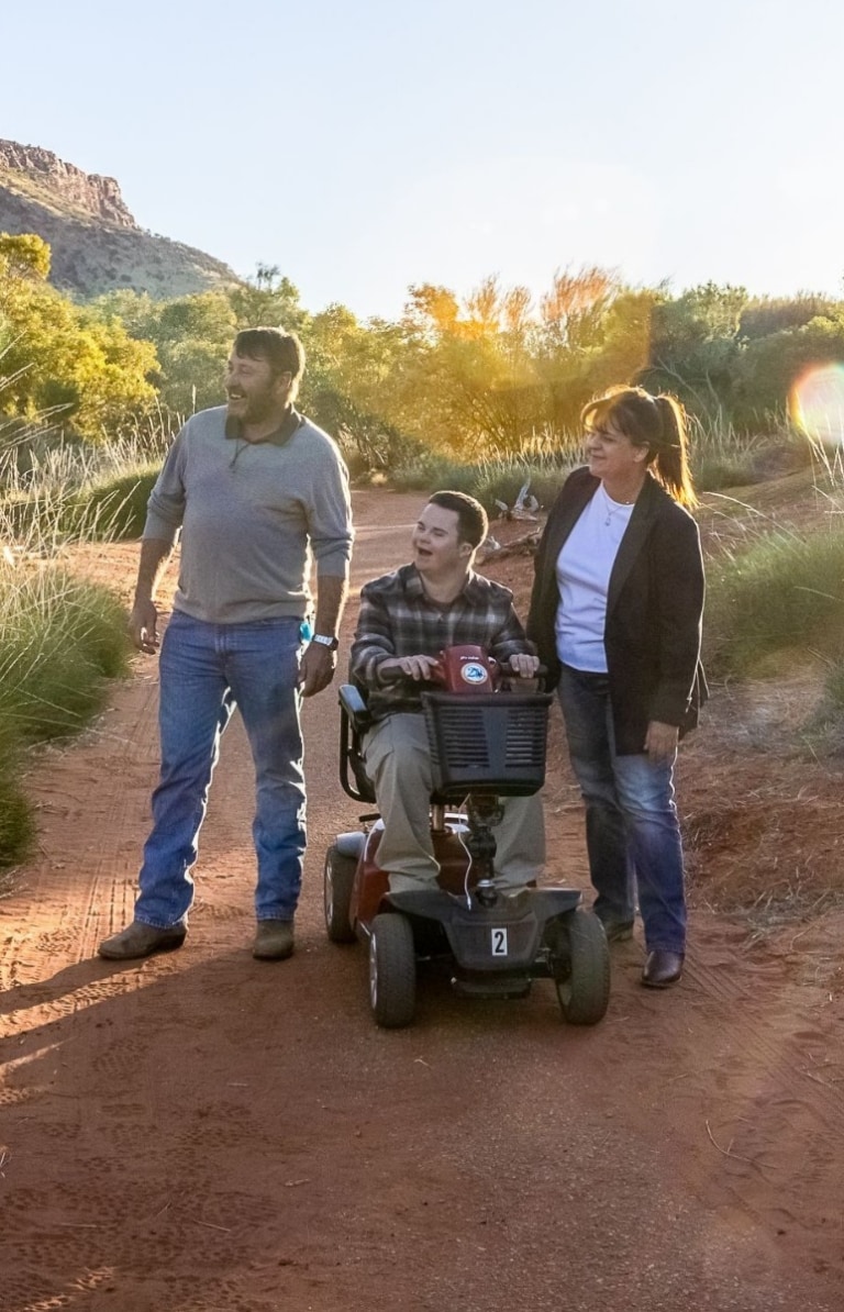 Ein E-Rollstuhlfahrer und zwei weitere Personen wandern zusammen durch das Grün und die rote Erde des Alice Springs Desert Park in Alice Springs, Northern Territory.