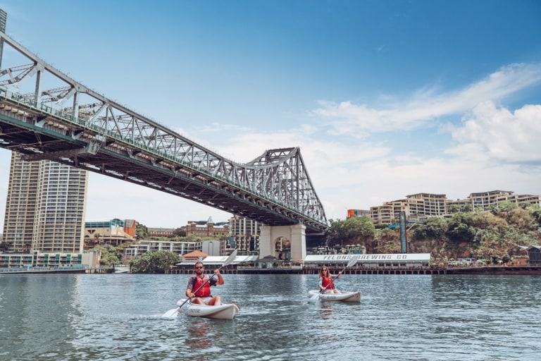 Ein Mann und eine Frau fahren mit Riverlife Kajak auf dem Brisbane River unter der Story Bridge mit den Howard Smith Wharves im Hintergrund in Brisbane, Queensland © Tourism and Events Queensland