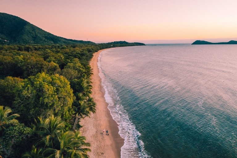 Luftaufnahme von zwei Personen, die bei Sonnenuntergang in Cairns am Sandstrand von Clifton Beach zwischen dem Regenwald und dem Ozean spazieren gehen, Queensland © Tourism and Events Queensland