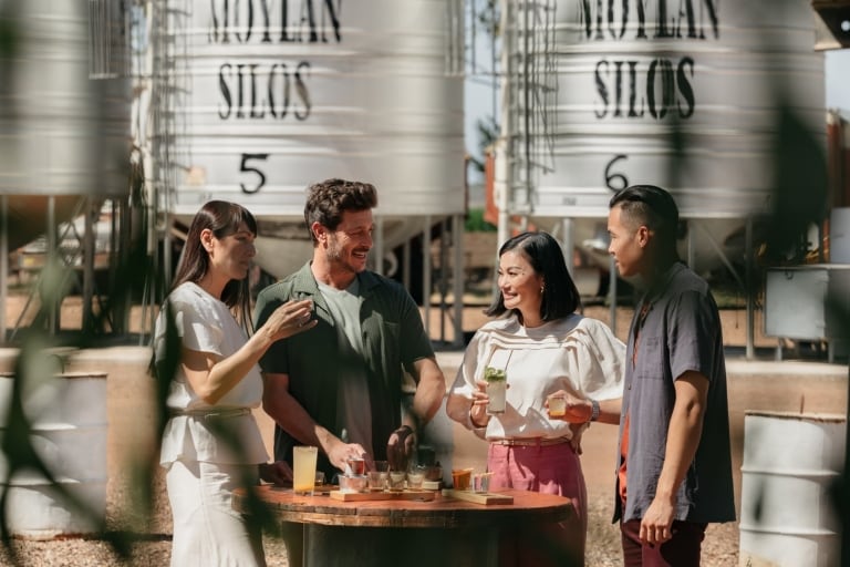 Four friends standing around a table outside, sipping drinks with silos in the background at the Hoochery Distillery Café, Kununurra, Western Australia © Tourism Western Australia