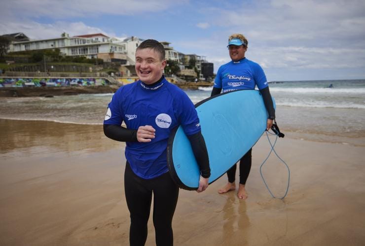 Homme en situation de handicap neurologique tenant une planche de surf sur le sable avec les moniteurs de Let's Go Surfing, Bondi Beach, Sydney, Nouvelle-Galles du Sud © Tourism Australia