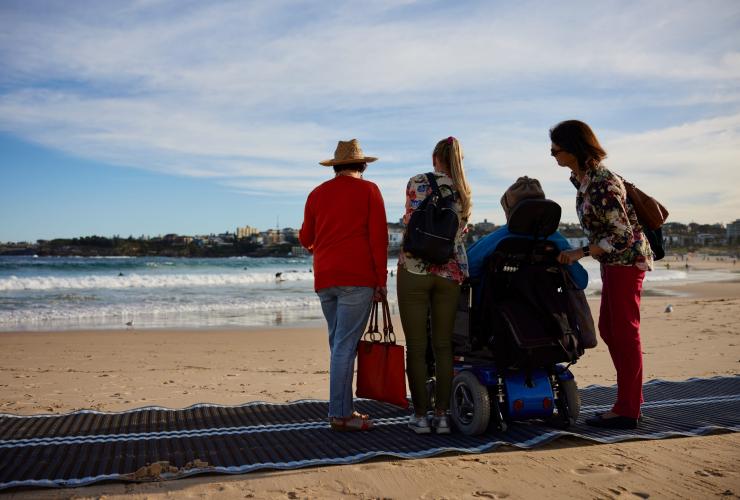 Homme en fauteuil sur un tapis d'accès plage avec sa famille à Bondi Beach avec Australia In Style, Sydney, Nouvelle-Galles du Sud © Destination NSW