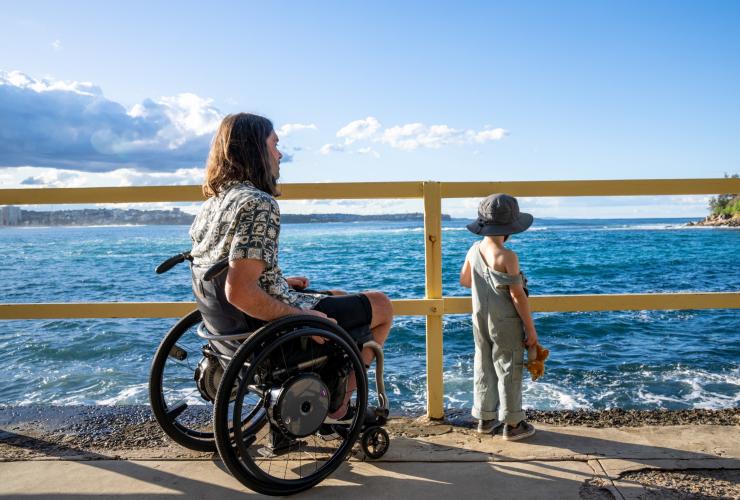 Seorang pria yang duduk di kursi roda memandang lautan bersama putranya di Manly, Sydney, New South Wales © Tourism Australia