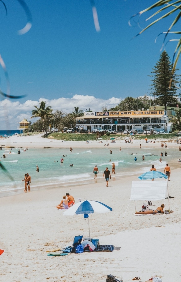 Orang-orang berbaring di pasir putih di bawah payung warna-warni sementara yang lainnya berenang di ombak yang tenang dan air biru jernih di Greenmount Beach di Coolangatta, Queensland © Tourism and Events Queensland