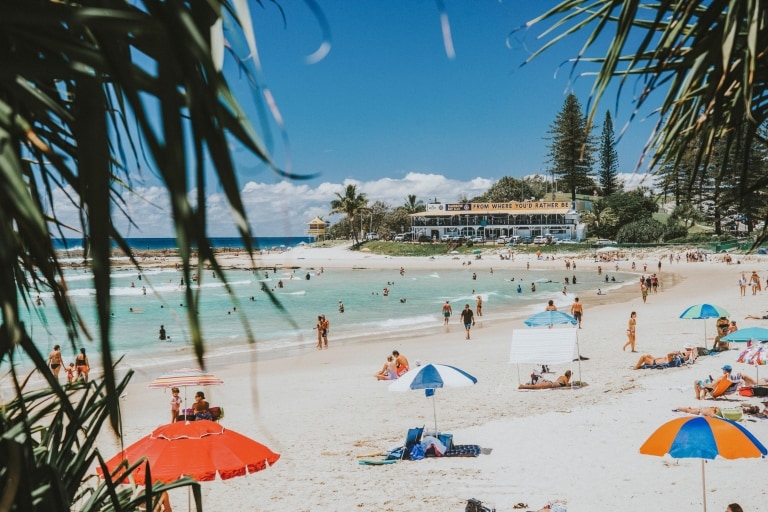Persone sdraiate sulla sabbia bianca sotto ombrelloni colorati e altre persone che nuotano tra le placide onde e nelle acque cristalline di Greenmount Beach a Coolangatta, Queensland © Tourism and Events Queensland