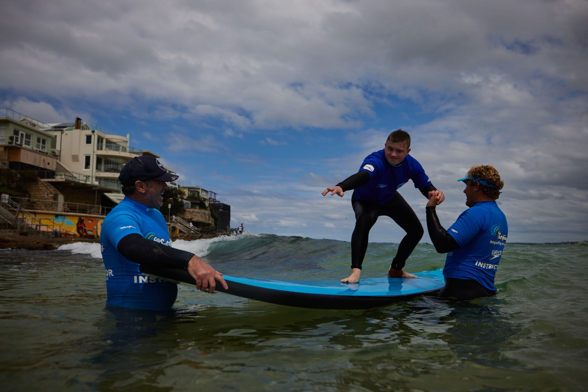 ニュー・サウス・ウェールズ州、シドニー、ボンダイ・ビーチ、レッツゴー・サーフィンのインストラクターの補助でサーフィンをする神経的多様性を持つ男性 © Tourism Australia