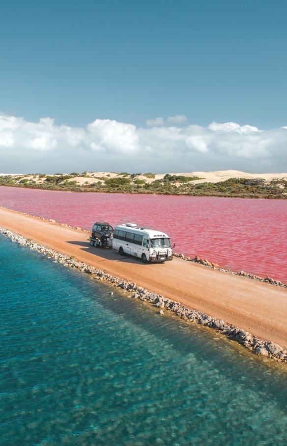 南オーストラリア州、エア半島に広がるピンク色のマクドネル湖とグリーン湖を結ぶ未舗装の道路を走り抜けるバン © Jaxon Foale
