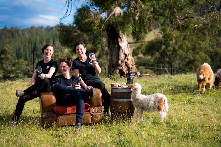 Drei Menschen lächeln in die Kamera, umgeben von Schafen und Weiden © Tourism Tasmania und Rob Burnett