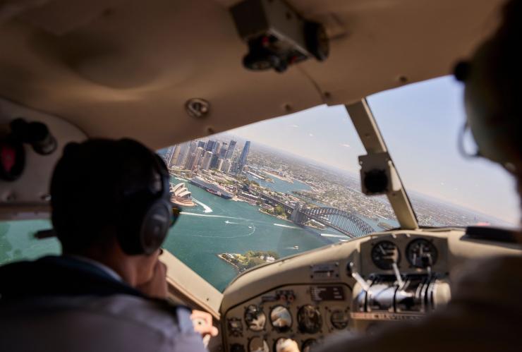 Pilot eines Wasserflugzeugs, der auf das Sydney Opera House, die Sydney Harbour Bridge und den glitzernden Ozean in Sydney blickt, New South Wales © Destination NSW
