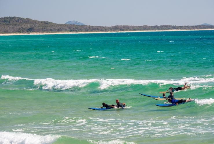 Mehrere Surfer liegen am Noosa Main Beach auf ihren Surfbrettern, während hinter ihnen eine Welle mit Schaum anrollt, Noosa, Queensland © Tourism Australia