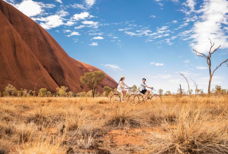 Two people cycling along a grassy plain with the towering red rock walls of Uluru behind them in Uluru-Kata Tjuta National Park, Northern Territory © Tourism NT