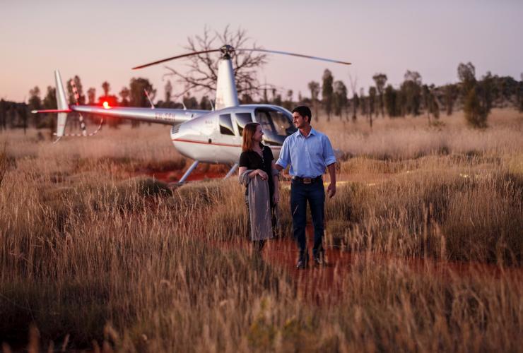 Pasangan berjalan melintasi tanah merah dan rerumputan dengan latar belakang helikopter di Longitude 131, Yulara, Northern Territory © Tourism Australia
