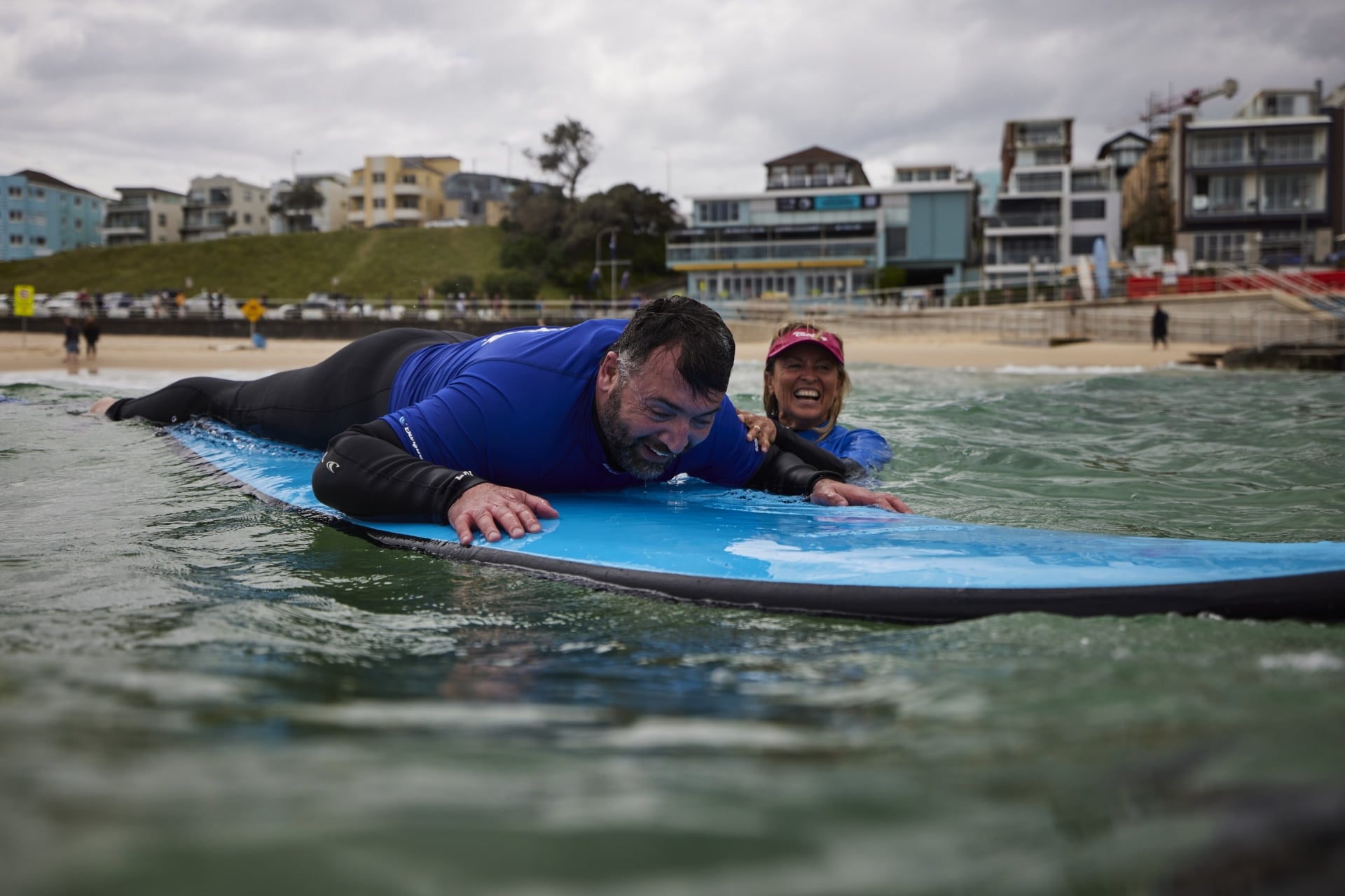Mann mit eingeschränktem Sehvermögen auf einem Surfbrett, der von einem Surflehrer von Let's Go Surfing geführt wird, Bondi Beach, New South Wales © Tourism Australia