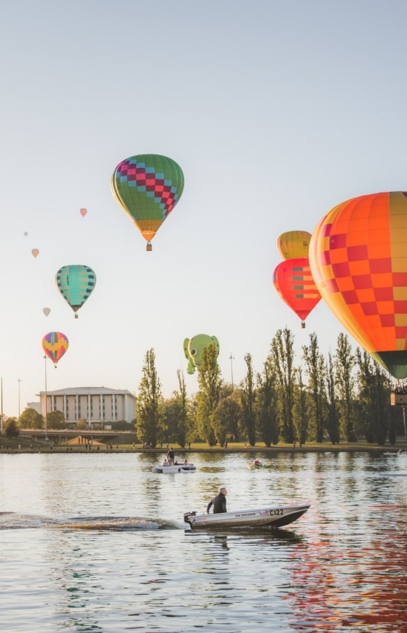 Heißluftballons schweben über dem ruhigen Wasser des Lake Burley Griffin, der mit Booten übersät ist, in Canberra, Australian Capital Territory © EventsACT