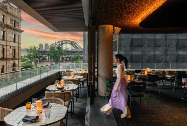 Eine Person spaziert während eines farbenprächtigen rosa Sonnenuntergangs über den Balkon eines Restaurants mit kerzenbeleuchteten Tischen und Blick auf eine große Stahlbogenbrücke. Penelope’s, Sydney, New South Wales © Penelope’s/Chad Konik