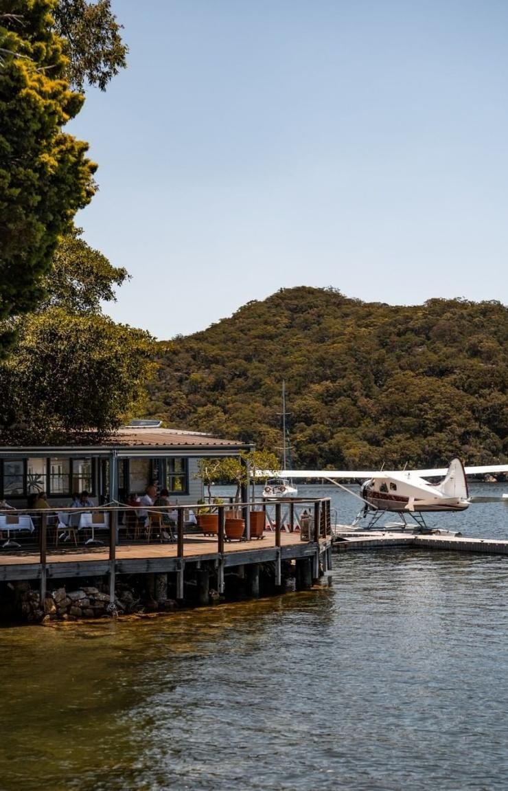 Gäste genießen ihr Essen in einem Restaurant am Flussufer, umgeben von Buschland, während ein Wasserflugzeug am Steg liegt, Cottage Point Inn, Cottage Point, Ku-ring-gai Chase National Park, New South Wales © Brett Stevens/Bennelong