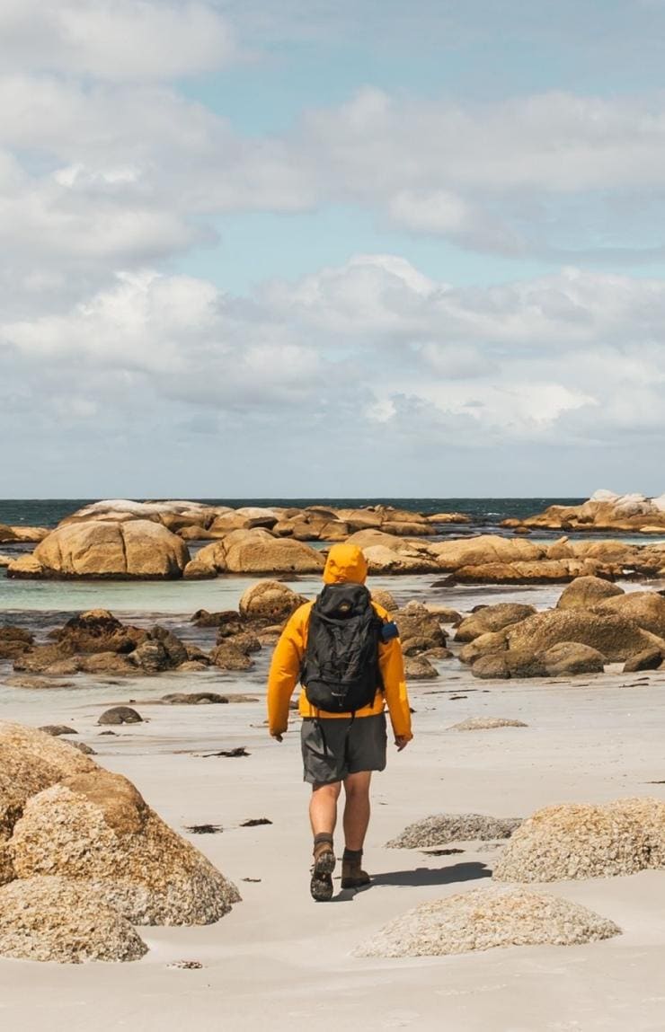 Eine Person spaziert entlang eines weißen Sandstrands mit großen, orangefarbenen Felsen 