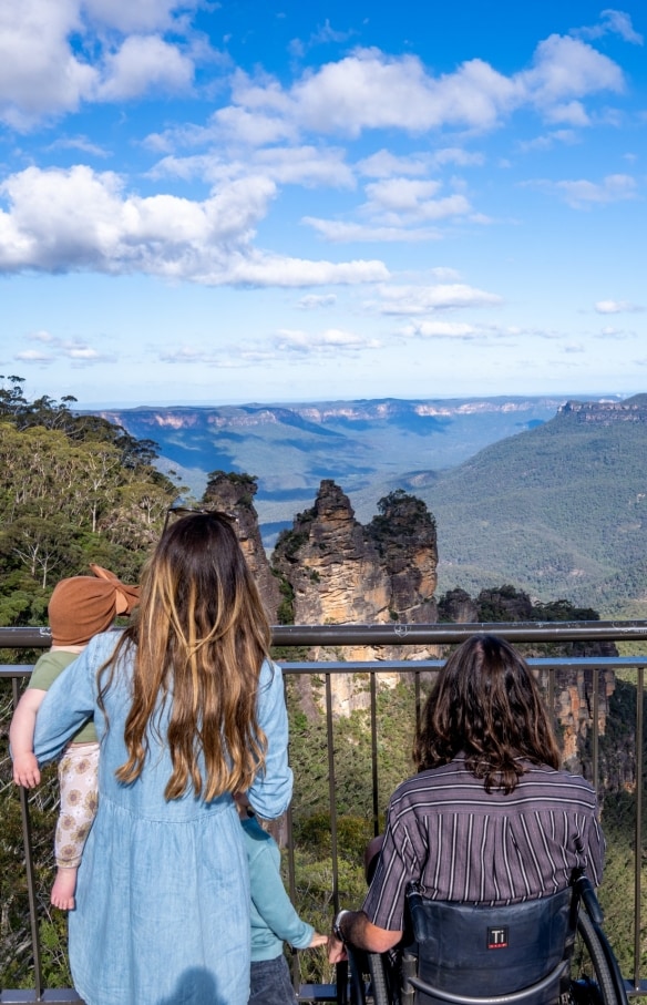 Ein Mann in einem Rollstuhl mit seiner Familie, die auf die Three Sisters im Blue Mountains National Park schauen, New South Wales © Tourism Australia