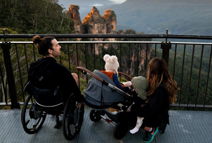 Ein Mann in einem Rollstuhl mit seiner Familie, die auf die Three Sisters in den Blue Mountains schauen, New South Wales © Tourism Australia