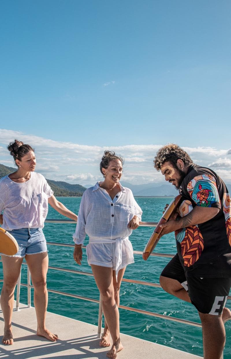 Vier lächelnde Personen auf einem Boot auf dem türkisfarbenen Meer, im Hintergrund eine üppig grüne Küste, während zwei Aboriginal Guides in traditioneller Bemalung Artefakte präsentieren, Dreamtime Dive and Snorkel, Great Barrier Reef, Cairns, Queensland © Tourism and Events Queensland