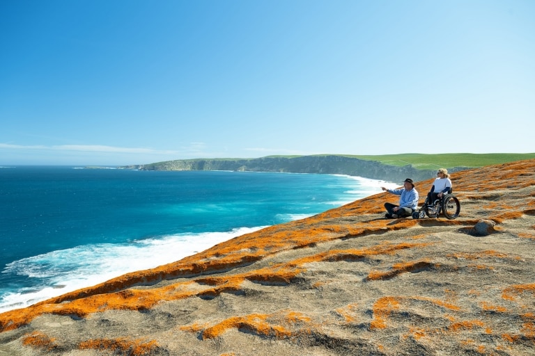 Eine Frau im Rollstuhl neben einem Tour-Guide, der auf den blauen Ozean zeigt, Kangaroo Island, Südaustralien © Tourism Australia