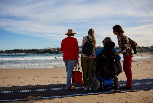 A group of people on beach matting atop a golden sand beach looking out towards the blue ocean with one person seated in a mobility device on Bondi Beach, Australia In Style, Sydney, New South Wales © Destination NSW