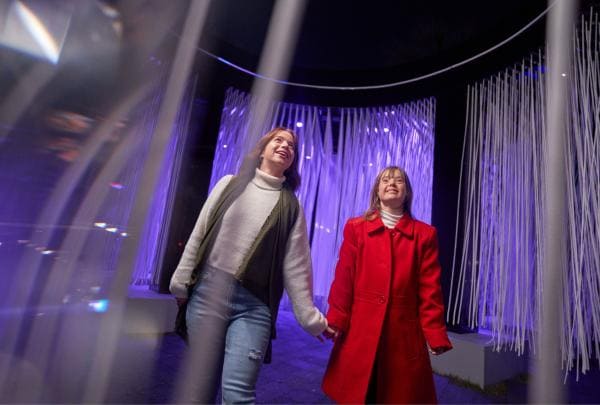 A person with neurodivergence holding hands with another person while walking through an art installation of hanging lights at Vivid Sydney 2024, New South Wales © Destination New South Wales