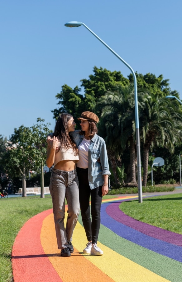 A couple with their arms around one another as they wander along a rainbow pathway through the green grass and trees of Prince Alfred Park, Surry Hills, New South Wales © Destination NSW