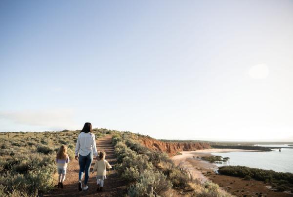 A woman and two children walking on a path bordered by bushland along a headland overlooking a calm blue waterway at Australian Arid Lands Botanic Garden, Port Augusta, South Australia © Tourism Australia / South Australian Tourism Commission    