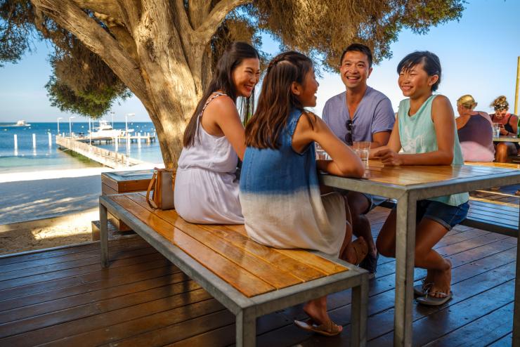 A family seated around an outdoor picnic table beneath a tree with the ocean in the background at Hotel Rottnest, Rottnest Island, Western Australia © Tourism Western Australia A family seated around an outdoor picnic table beneath a tree with the ocean in the background at Hotel Rottnest, Rottnest Island, Western Australia © Tourism Western Australia