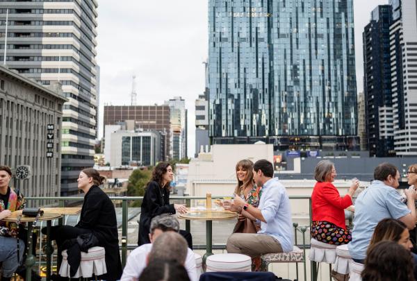 Three friends smiling while enjoying food and cocktails on a rooftop bar with a city skyline behind them at HER Bar, Melbourne, Victoria © Visit Victoria