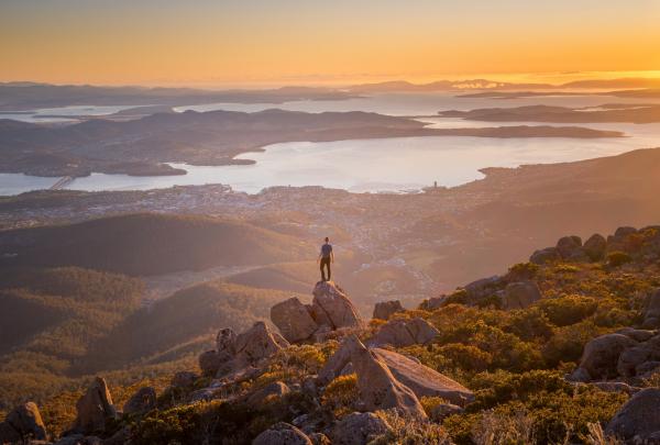 A man standing on a rock at the peak of a mountain overlooking a landscape of hills, waterways and a city on Mount Wellington/kunanyi, Hobart, Tasmania © Justin Hyde