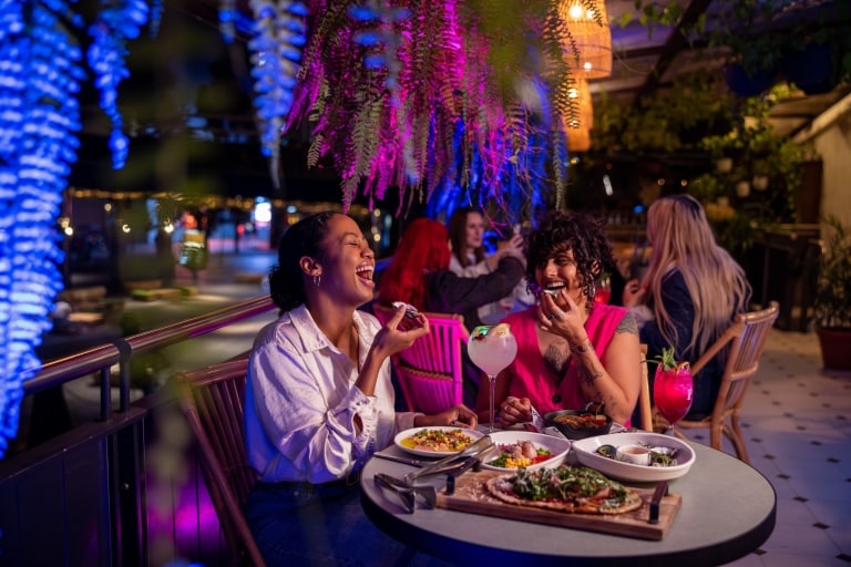Two women laugh as they enjoy plates of food and cocktails at Pumphouse Bar and Restaurant in Sydney, New South Wales © Destination NSW