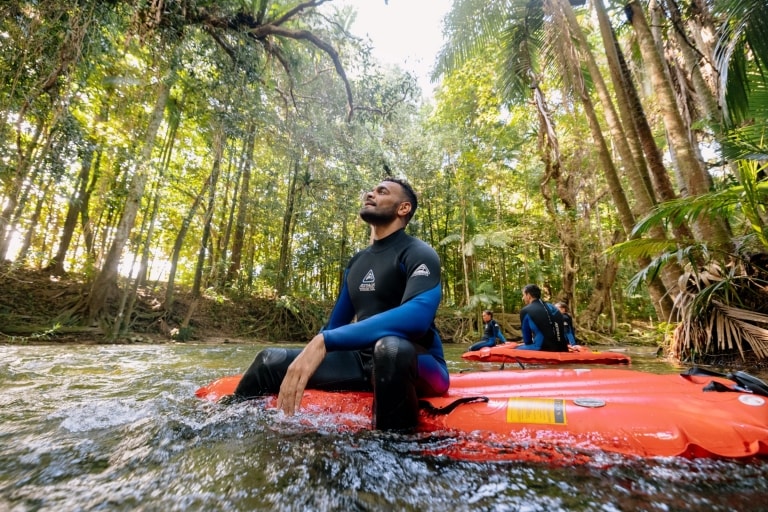A man sits on a floating raft in a river and gazes up at a green rainforest canopy with Back Country Bliss Adventure in the Daintree Rainforest, Queensland © Tourism Australia