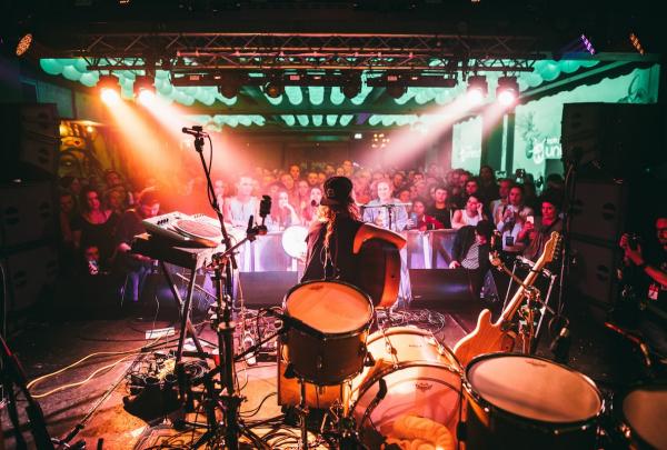 A performer sitting with a guitar on a stage behind a microphone with a large audience facing the camera beneath colourful lights during Bigsound, Fortitude Valley, Brisbane, Queensland © Stills in Time
