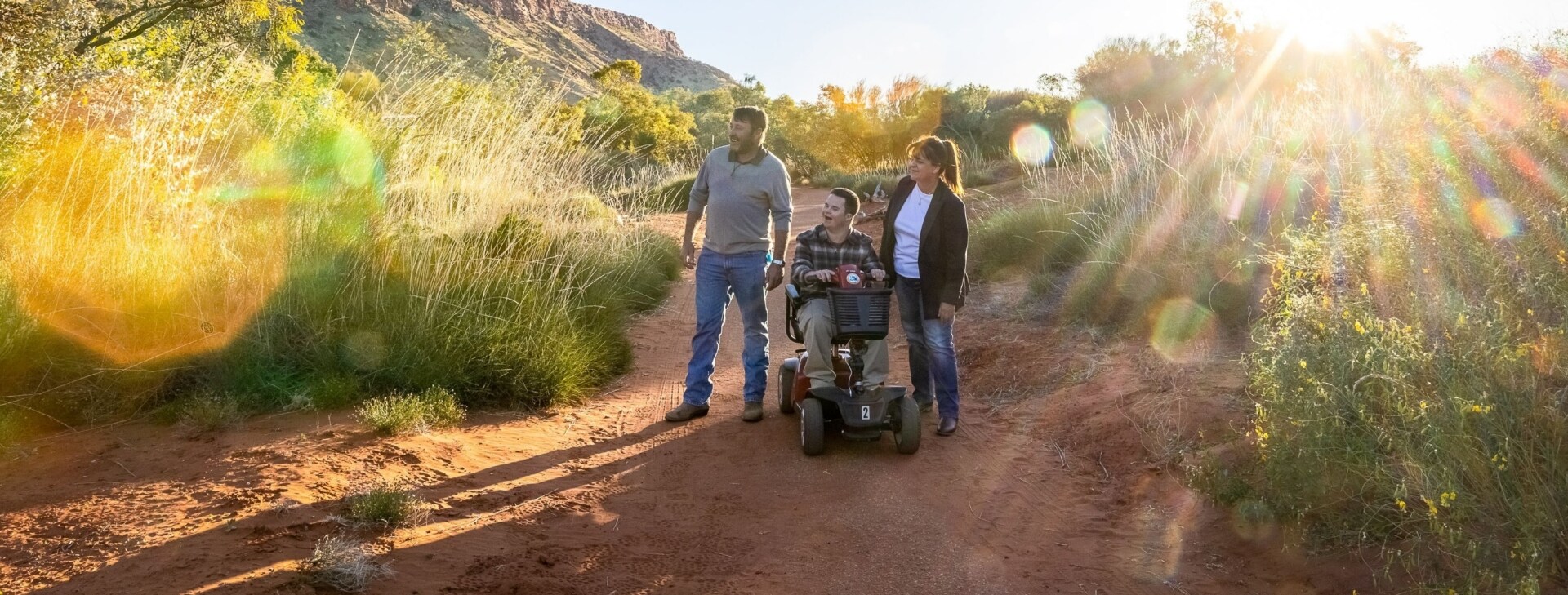 Man seated on a mobility device with another man and woman, wandering among the red dirt and greenery of Alice Springs Desert Park with the sun in the background in Alice Springs, Northern Territory © Tourism NT/Helen Orr