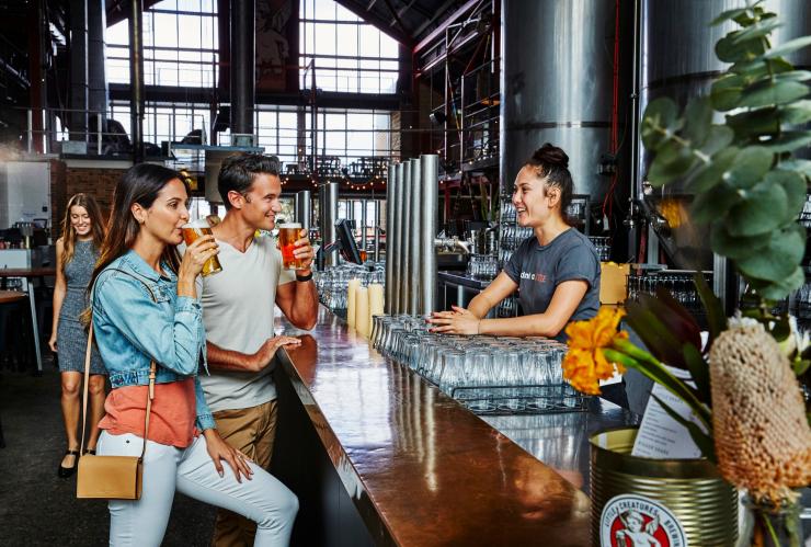 A bartender smiles while making conversation with two customers as they sip on schooners of beer in an industrial-styled venue at Little Creatures Brewery, Fremantle, Western Australia © Tourism Western Australia