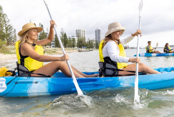 Two friends paddling in a blue kayak through clear water with a city skyline in the background during a tour with Australian Kayaking Adventures, Gold Coast, Queensland © Tourism Australia