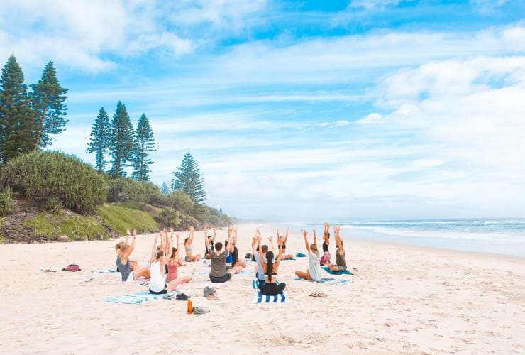 A group of people sitting on towels with their hands raised in the air on a white sand beach with pine trees to their left and the ocean to their right during a yoga class with Wake Up! Byron Bay Hostel, Byron Bay, New South Wales © Wake Up! Byron Bay