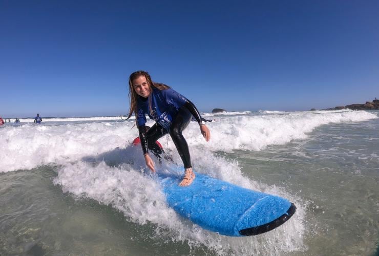 A person smiling while standing up on a blue surf board as an instructor pushes them onto a gentle wave with Margaret River Surf School, Redgate Beach, Margaret River, Western Australia © Tourism Australia