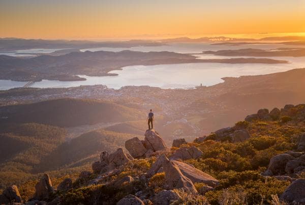 A man standing on a rock at the peak of a mountain overlooking a landscape of hills, waterways and a city on Mount Wellington/kunanyi, Hobart, Tasmania © Justin Hyde