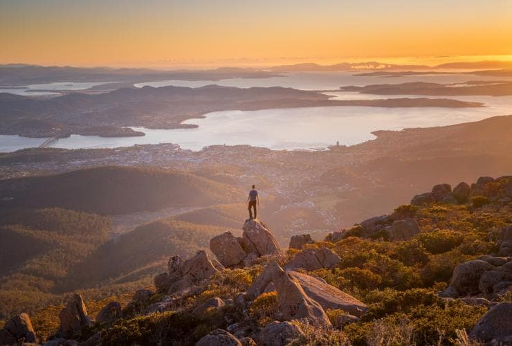 A man standing on a rock at the peak of a mountain overlooking a landscape of hills, waterways and a city on Mount Wellington/kunanyi, Hobart, Tasmania © Justin Hyde