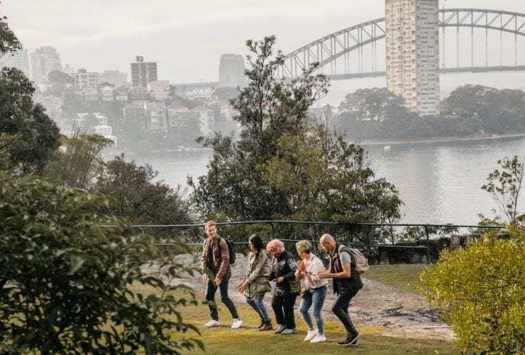 A group of people dancing with an Aboriginal tour guide among a leafy park with the Sydney Harbour Bridge in the background during a tour with Dreamtime Southern X, Sydney, New South Wales © Destination NSW