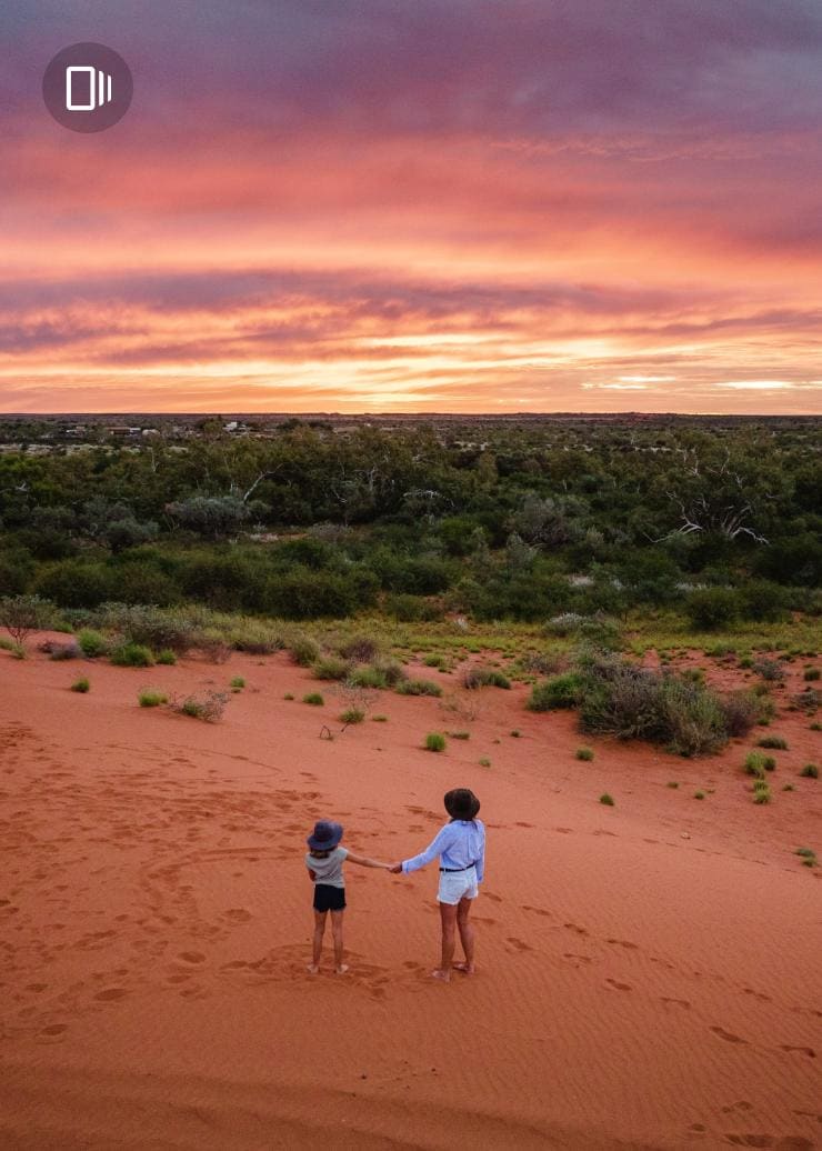 Aerial view over a mother and daughter standing hand-in-hand on a red sand landscape overlooking green bushland beneath a pink sunset, Western Australia © Tourism Australia