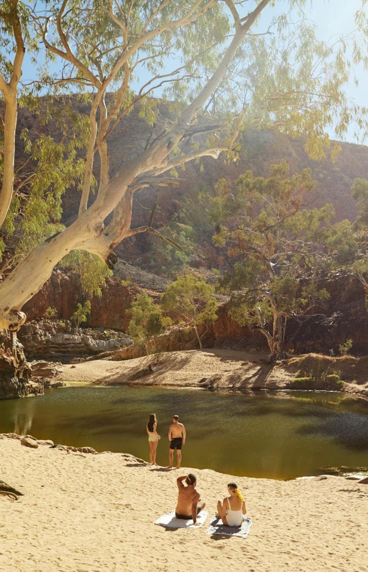 A group of friends relaxing at an outback waterhole surrounded by red rocky walls, Ormiston Gorge, West MacDonnell Ranges, Northern Territory © Tourism NT/Matt Cherubino 