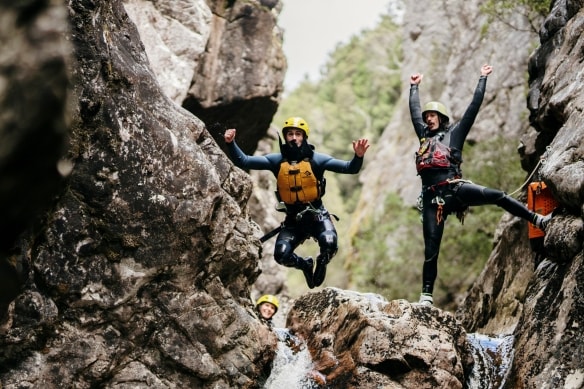 Two people wearing helments at life vests are seen at the top of a small waterfall as one of them jumps from the rock while the other cheers behind them with Cradle Mountain Canyons, Cradle Mountain, Tasmania © Tourism Australia