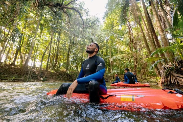 A man sits on a floating raft in a river and gazes up at a green rainforest canopy with Back Country Bliss Adventure in the Daintree Rainforest, Queensland © Tourism Australia