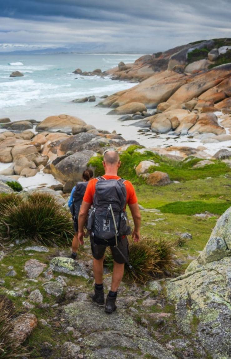 A group of hikers make their way down a rocky path toward a sandy beach with bright orange rocks, Bay of Fires Signature Walk, East Coast Tasmania, Tasmania © Tourism Australia