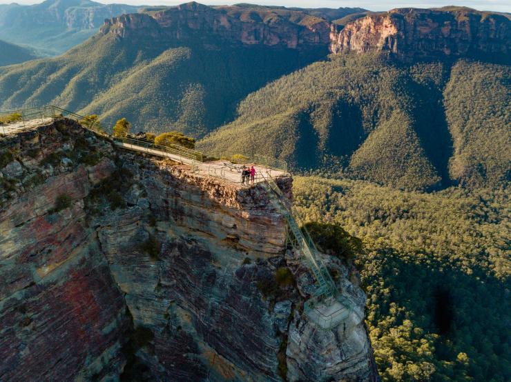 Aerial view over three people standing on a cliff-top lookout overlooking a lush green valley, Pulpit Rock Lookout, Blue Mountains, New South Wales © Destination NSW / The Travel Intern